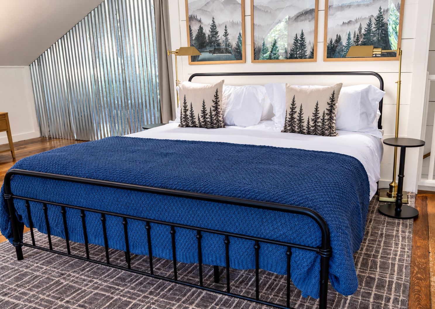 Cozy bedroom with black iron bed frame, navy blue quilt, white pillows, and mountain forest wall art above headboard.