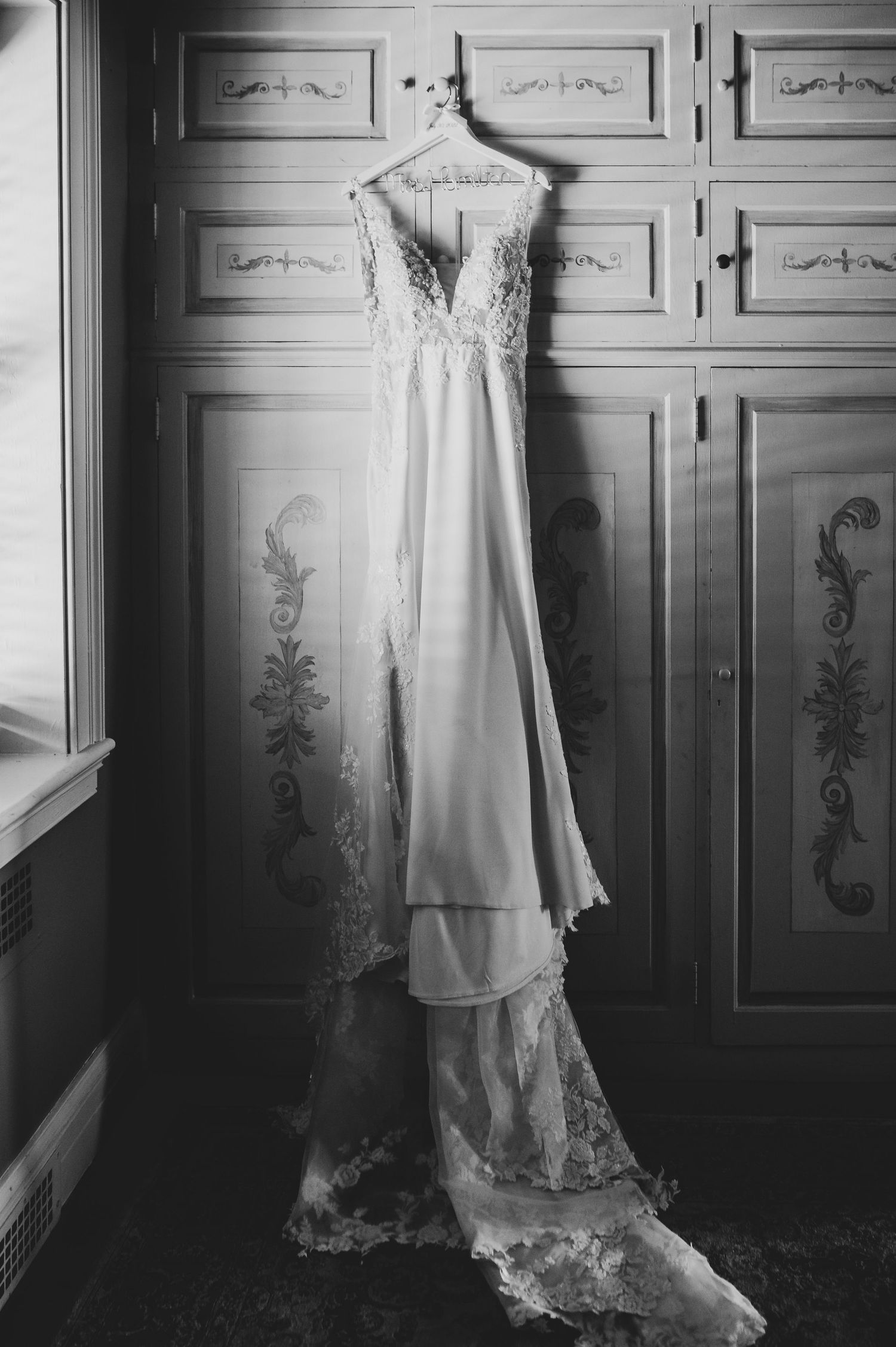 Black and white photo of an elegant lace wedding gown hanging on a hanger against ornate wooden cabinet doors.