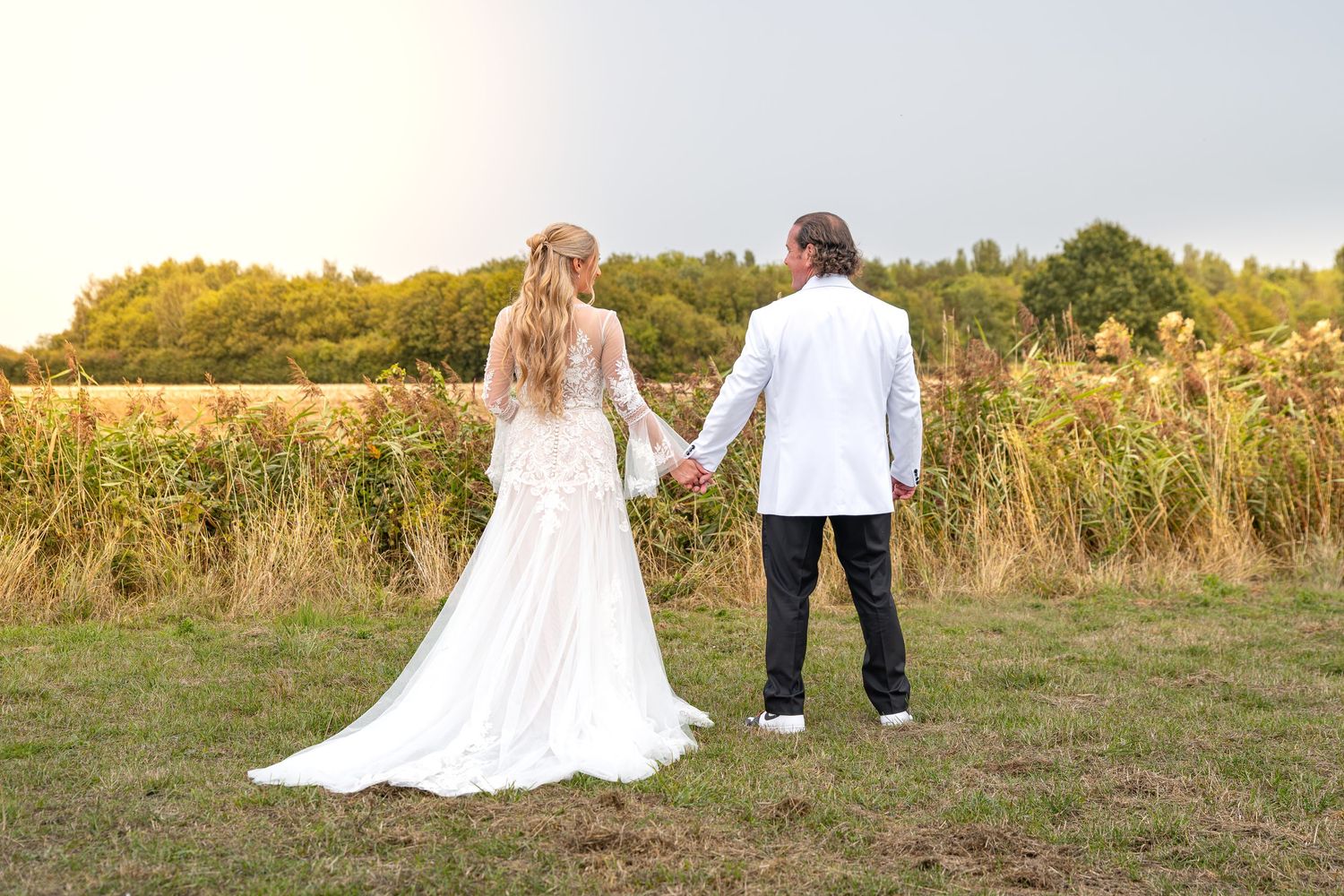Bride and groom holding hands facing a sunlit field and trees on their wedding day.Yorkshire, Sheffield, Lincolnshire, UK