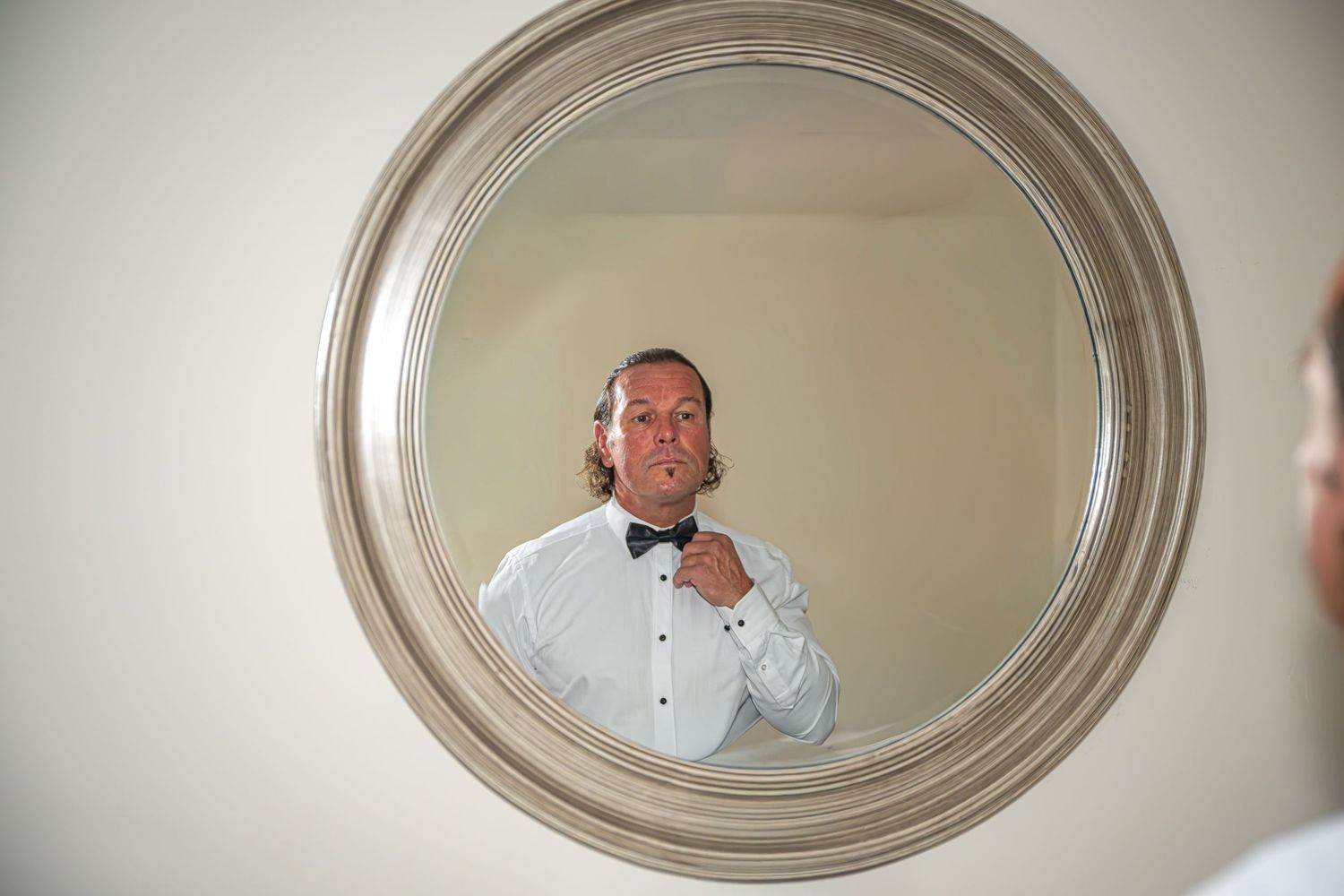 Photo of a groom adjusting his bow tie while looking at his reflection in a round silver mirror. Yorkshire, Sheffield, Lincolnshire, UK