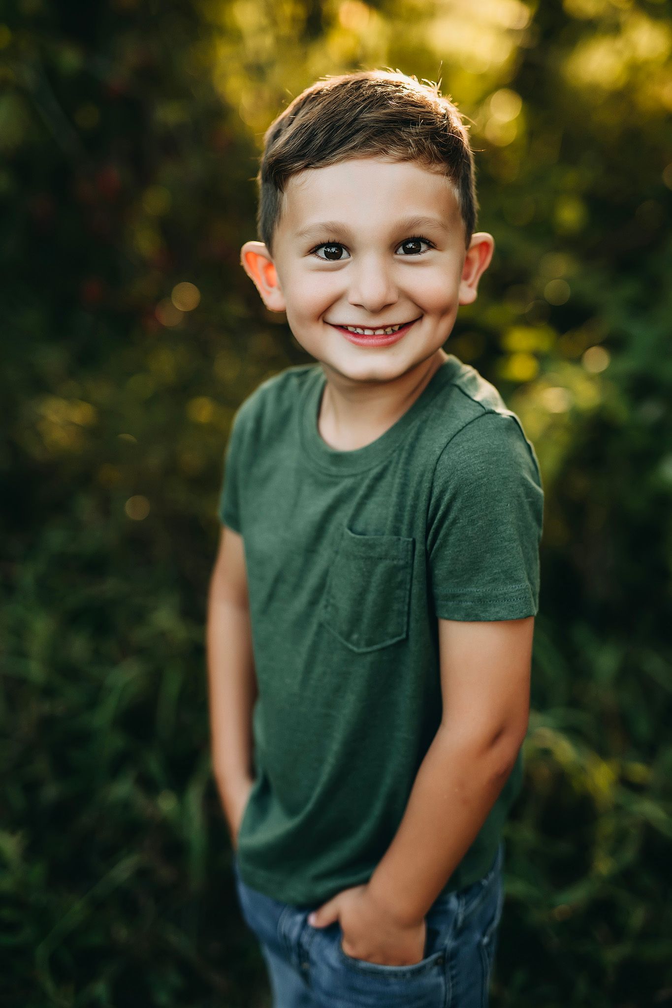 Smiling young boy in green t-shirt standing outdoors with lush green foliage background.