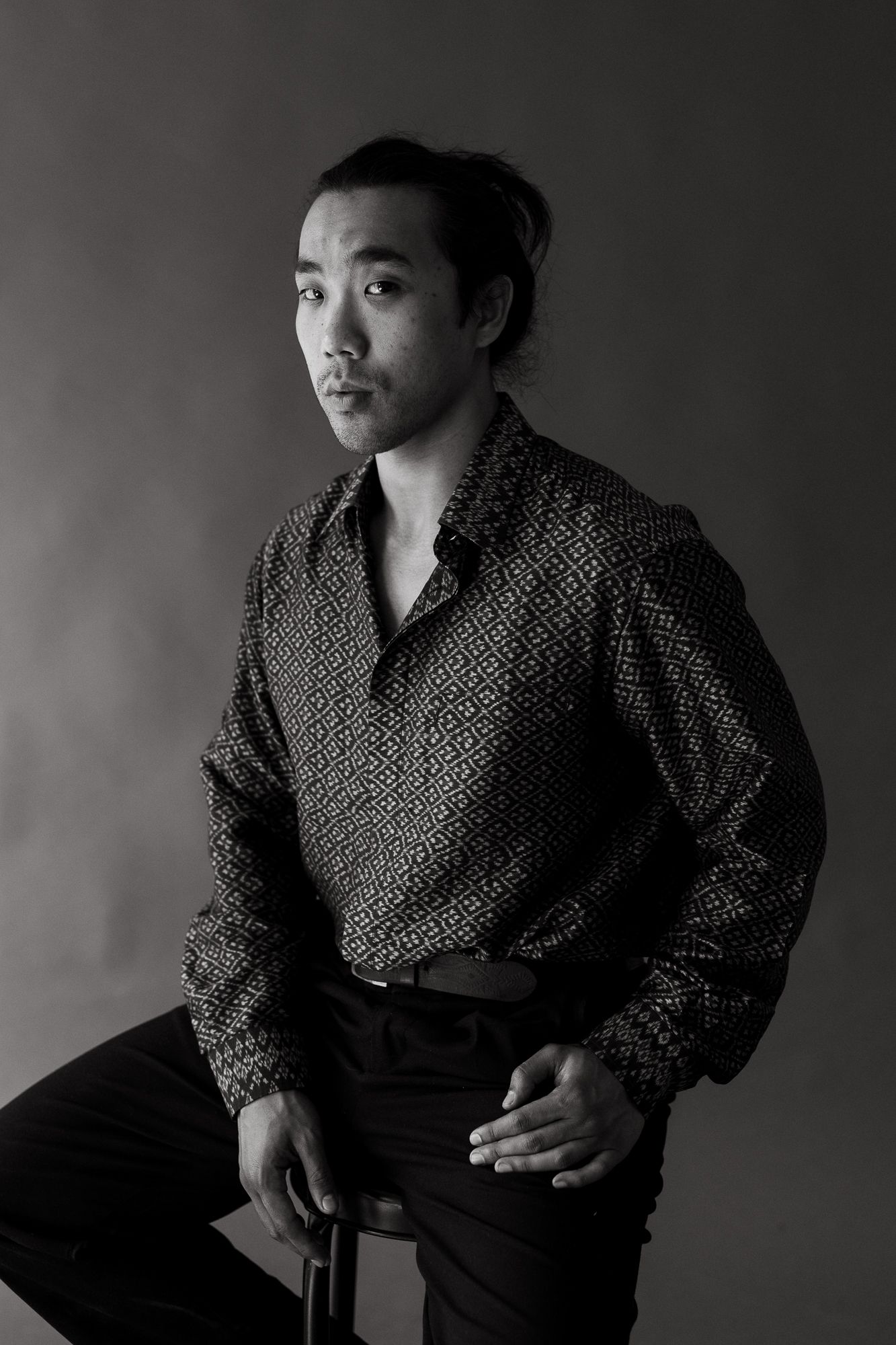 Black and white studio portrait of a young Asian man in a patterned shirt, seated and gazing thoughtfully to the side.