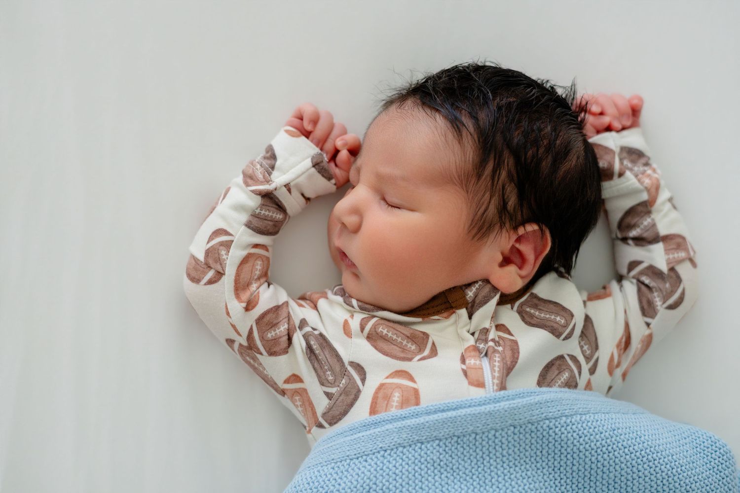 Peaceful newborn baby sleeping on back in patterned onesie with arms raised, wrapped in soft blue blanket.