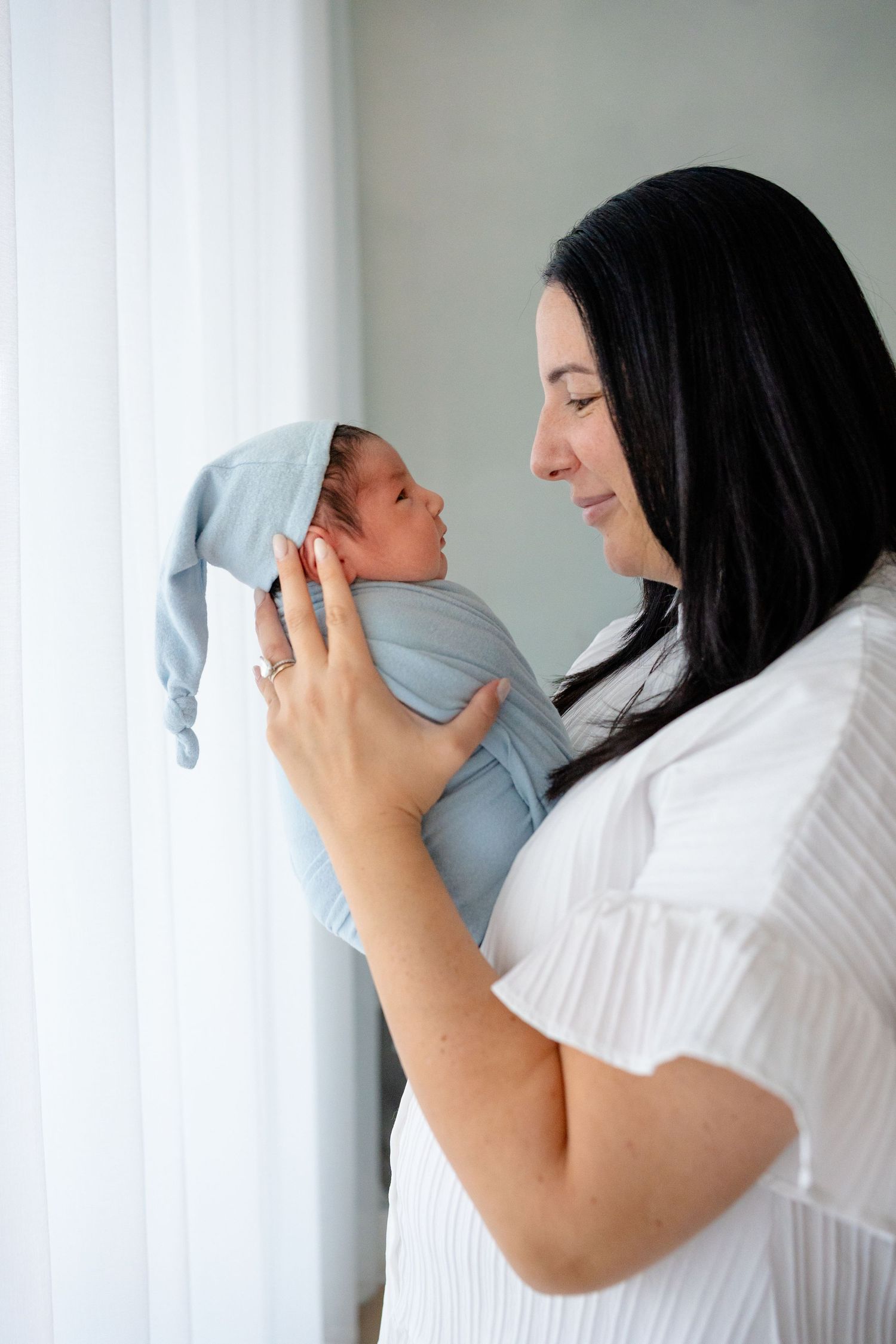 A mother tenderly holds her newborn baby in a blue hat and swaddle near a bright window.