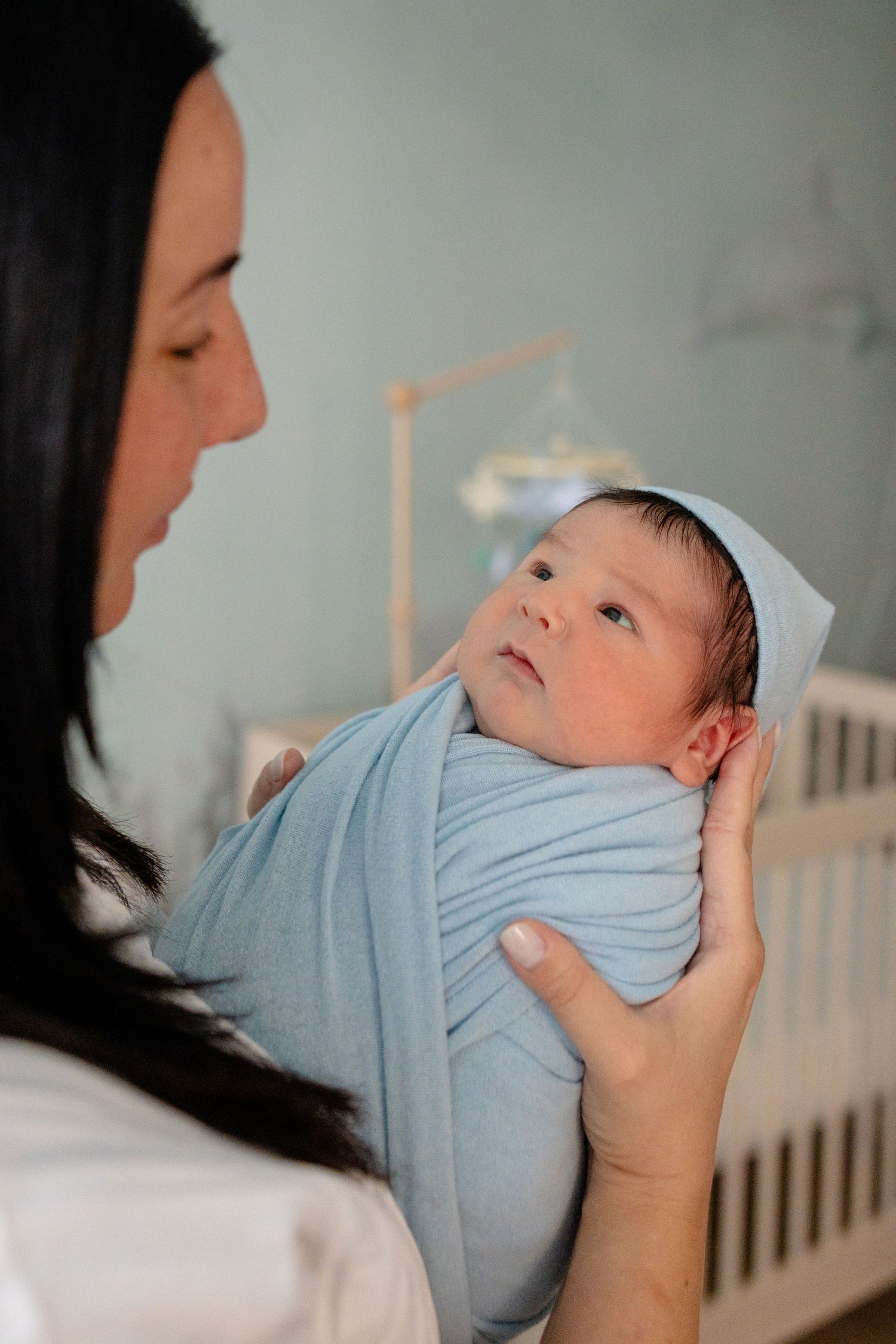 A mother tenderly holds her swaddled newborn baby in a blue wrap inside a nursery room.