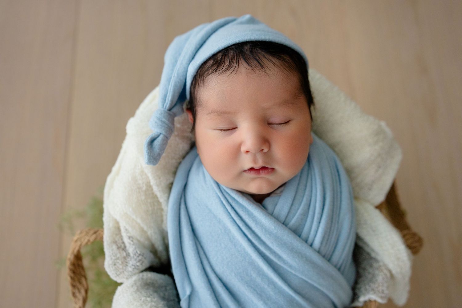 Newborn baby sleeping peacefully wrapped in a blue swaddle and matching knotted hat in a basket.