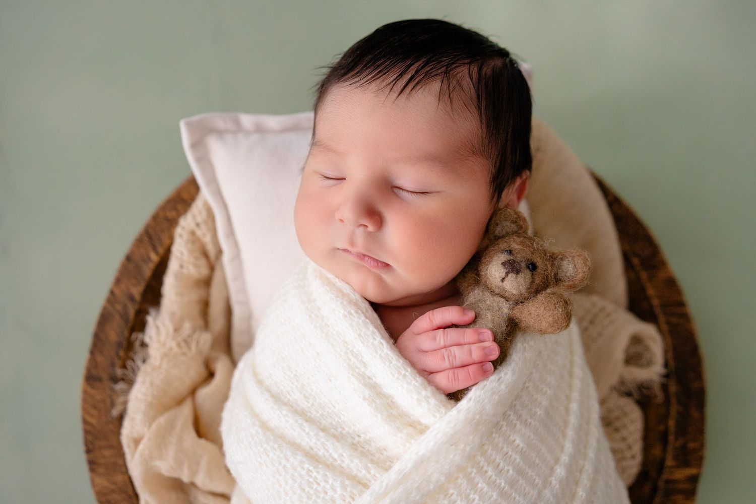 Newborn baby sleeping peacefully in a basket, swaddled in white wrap, holding a tiny teddy bear.