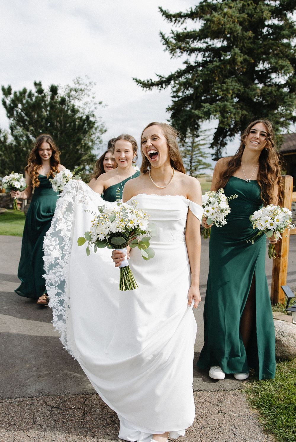 Bride in white off-shoulder gown laughs joyfully with bridesmaids in emerald green dresses holding white floral bouquets.