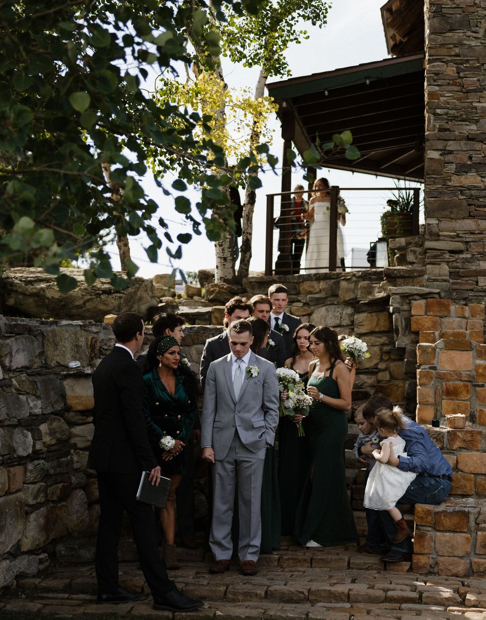 Wedding party descends rustic stone steps outdoors, groom in gray suit with bridesmaids in emerald green dresses.