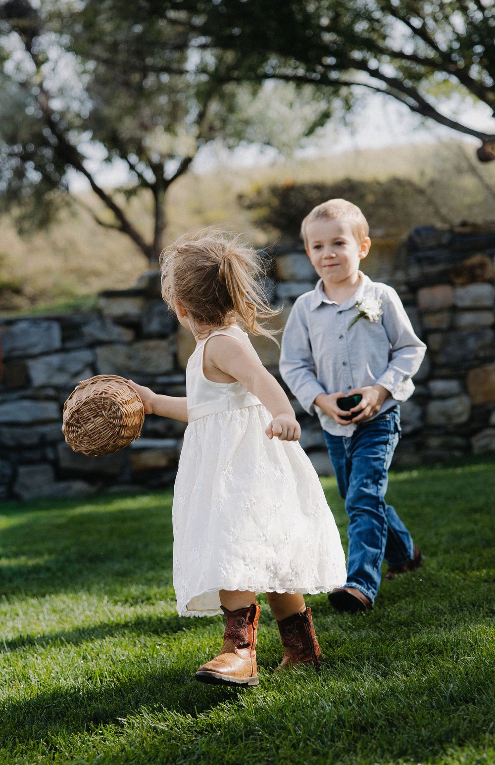 Flower girl in white dress and cowboy boots walks with ring bearer on green grass at outdoor wedding.
