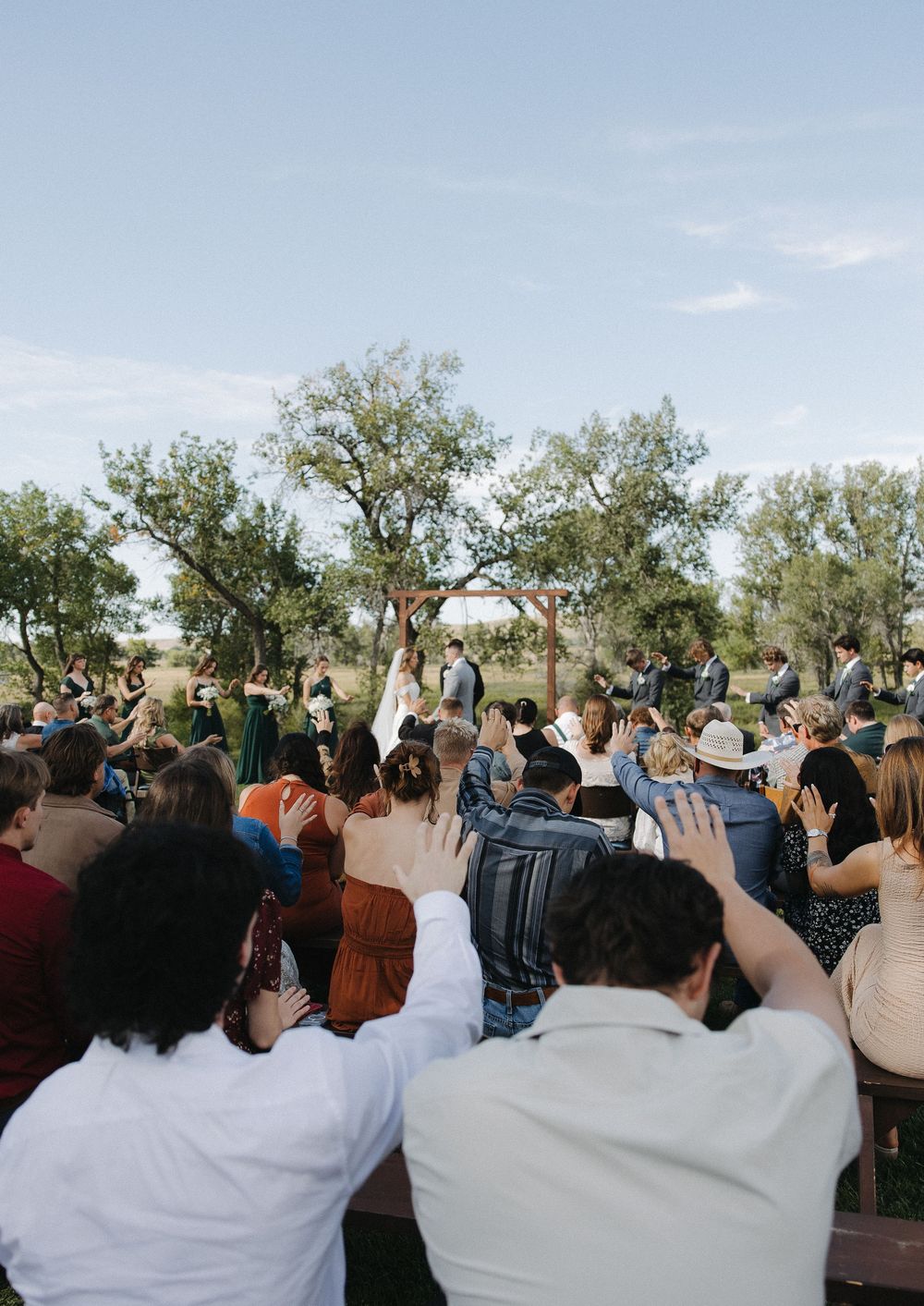Outdoor wedding ceremony with guests seated on benches, couple at altar under wooden arch surrounded by trees on a sunny day.