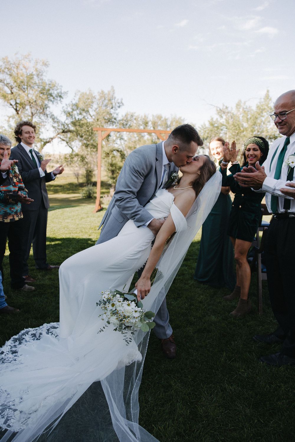 Groom dips bride for a kiss at outdoor wedding ceremony while guests cheer, bride holding white bouquet with veil flowing.