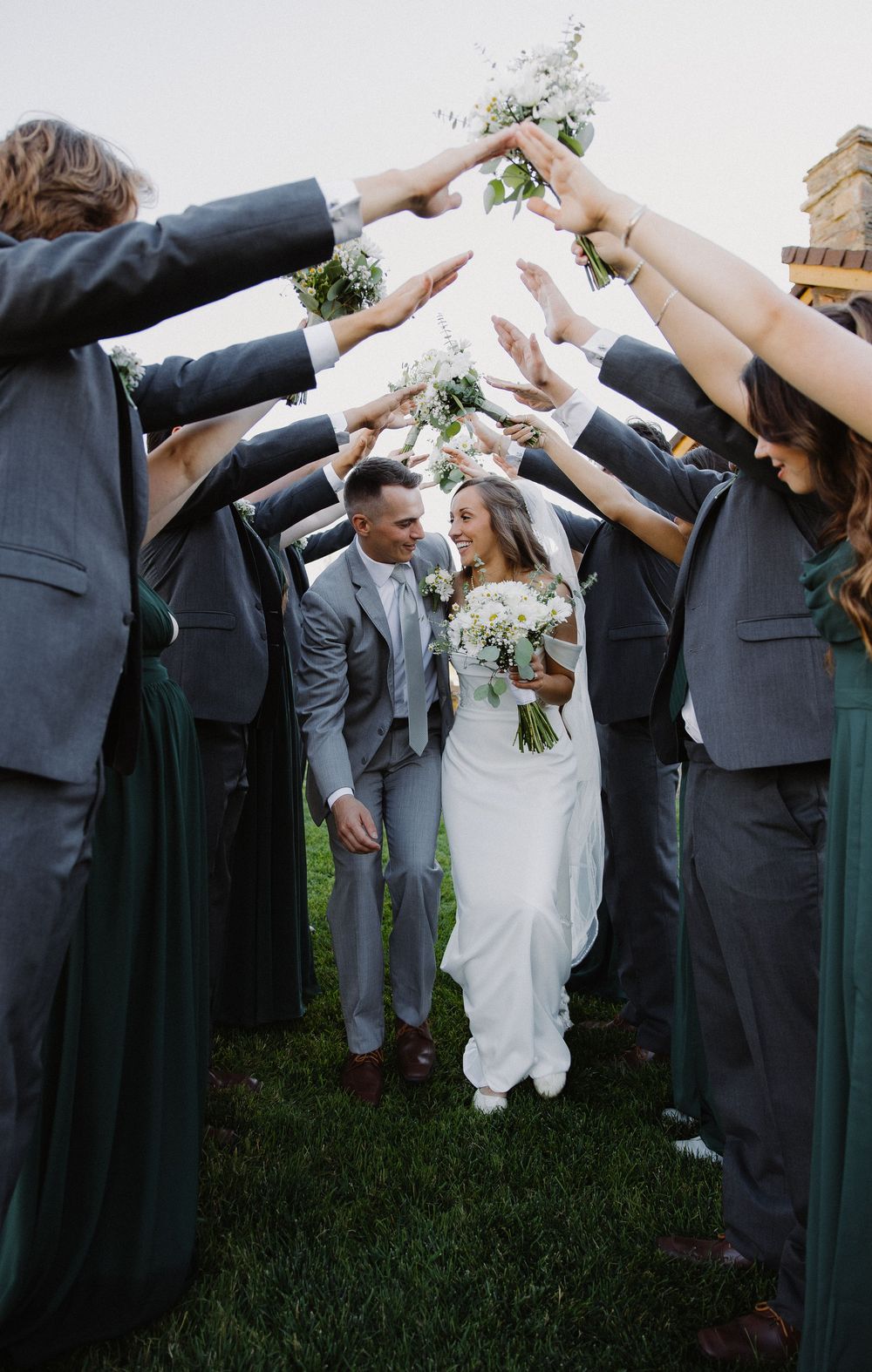 Bride and groom walk through an arch of raised hands formed by wedding party members in green and gray attire.