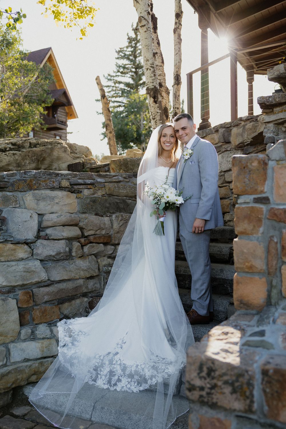 Bride in white gown with veil and groom in gray suit pose on stone steps surrounded by trees on their wedding day.