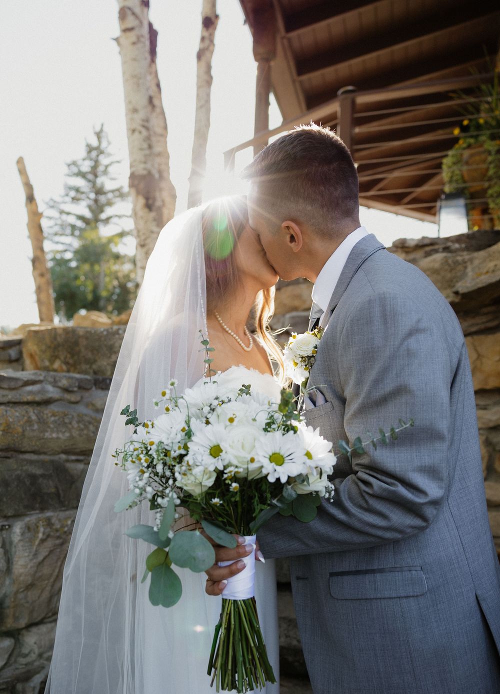 Bride and groom share a romantic kiss, sun flare glowing behind them, bride holding white daisy bouquet in wedding attire.