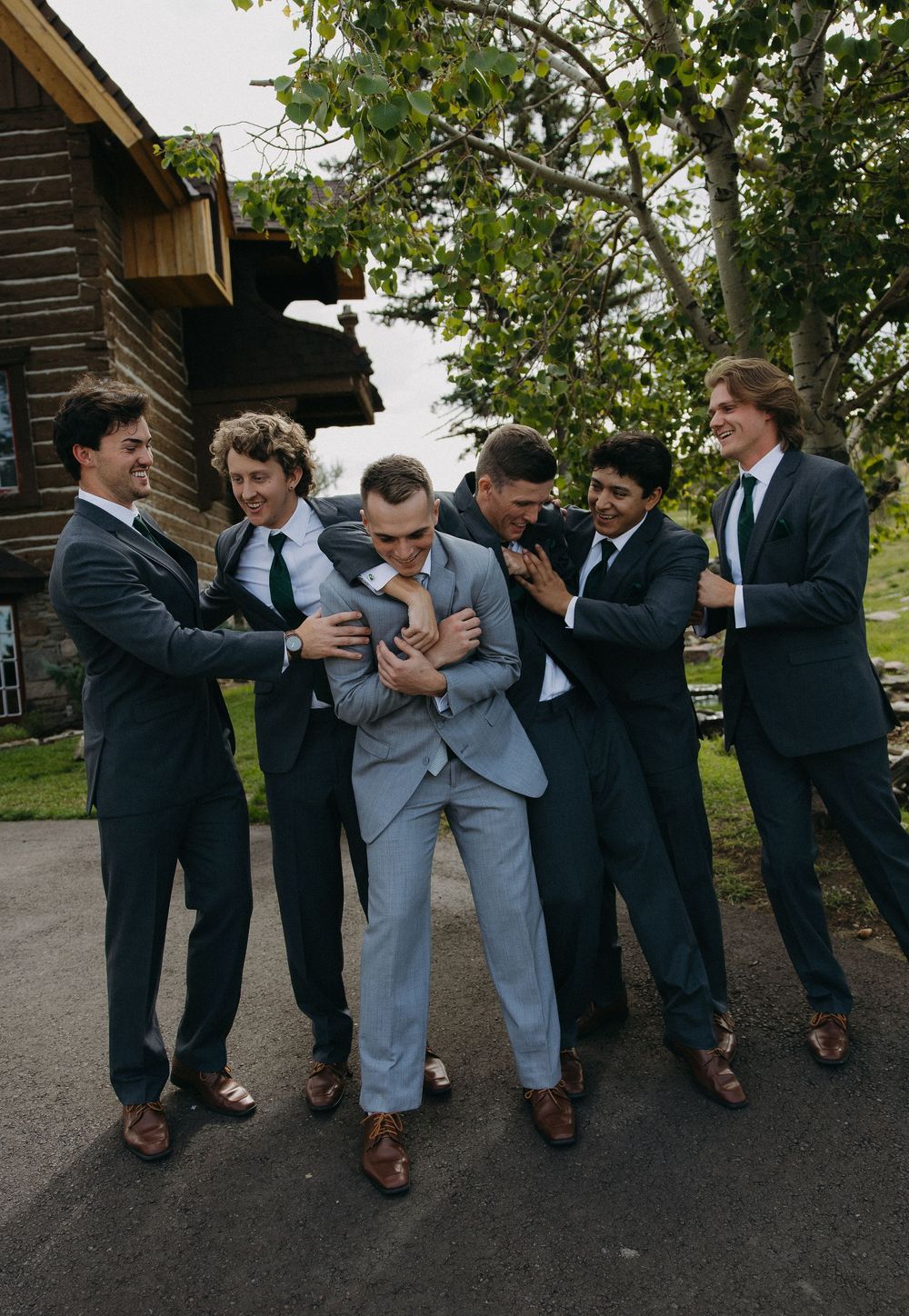 Groomsmen in dark suits laughing and playfully adjusting the groom's light gray suit outdoors near a rustic wooden building.