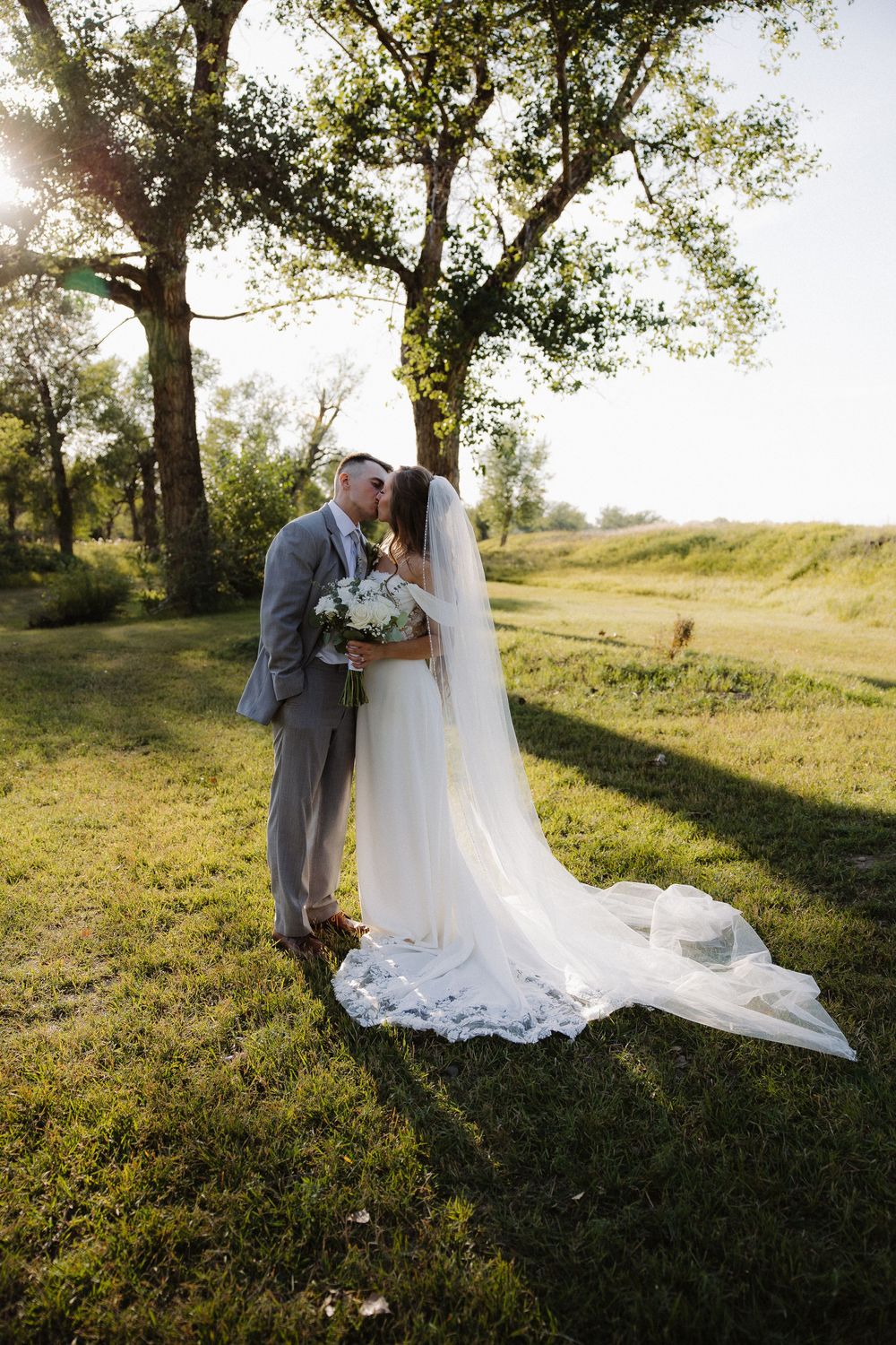 Bride and groom sharing a kiss outdoors under a tree, bride wearing a flowing white gown with long lace veil.