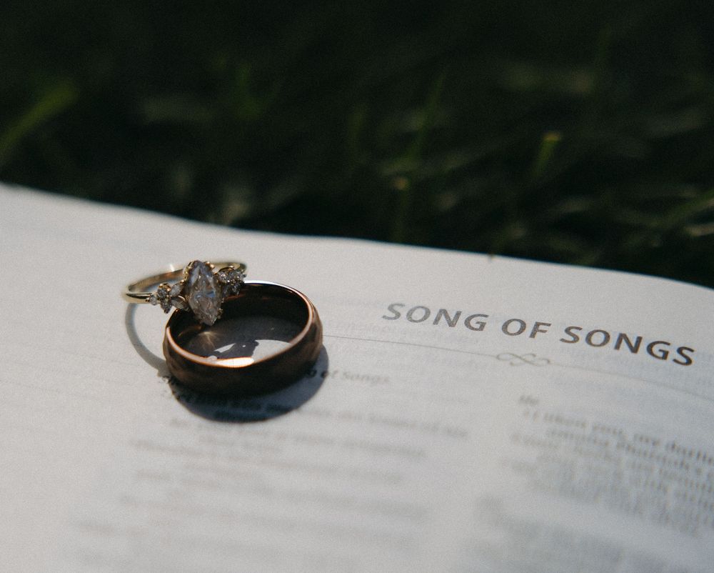 Wedding rings resting on an open Bible turned to the Song of Songs passage on a grassy background.