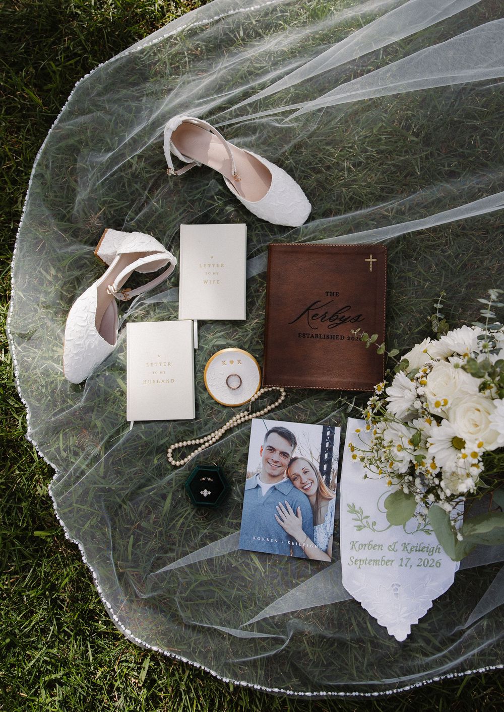 Elegant wedding flat lay featuring bridal shoes, veil, bouquet, rings, and invitation on green grass.