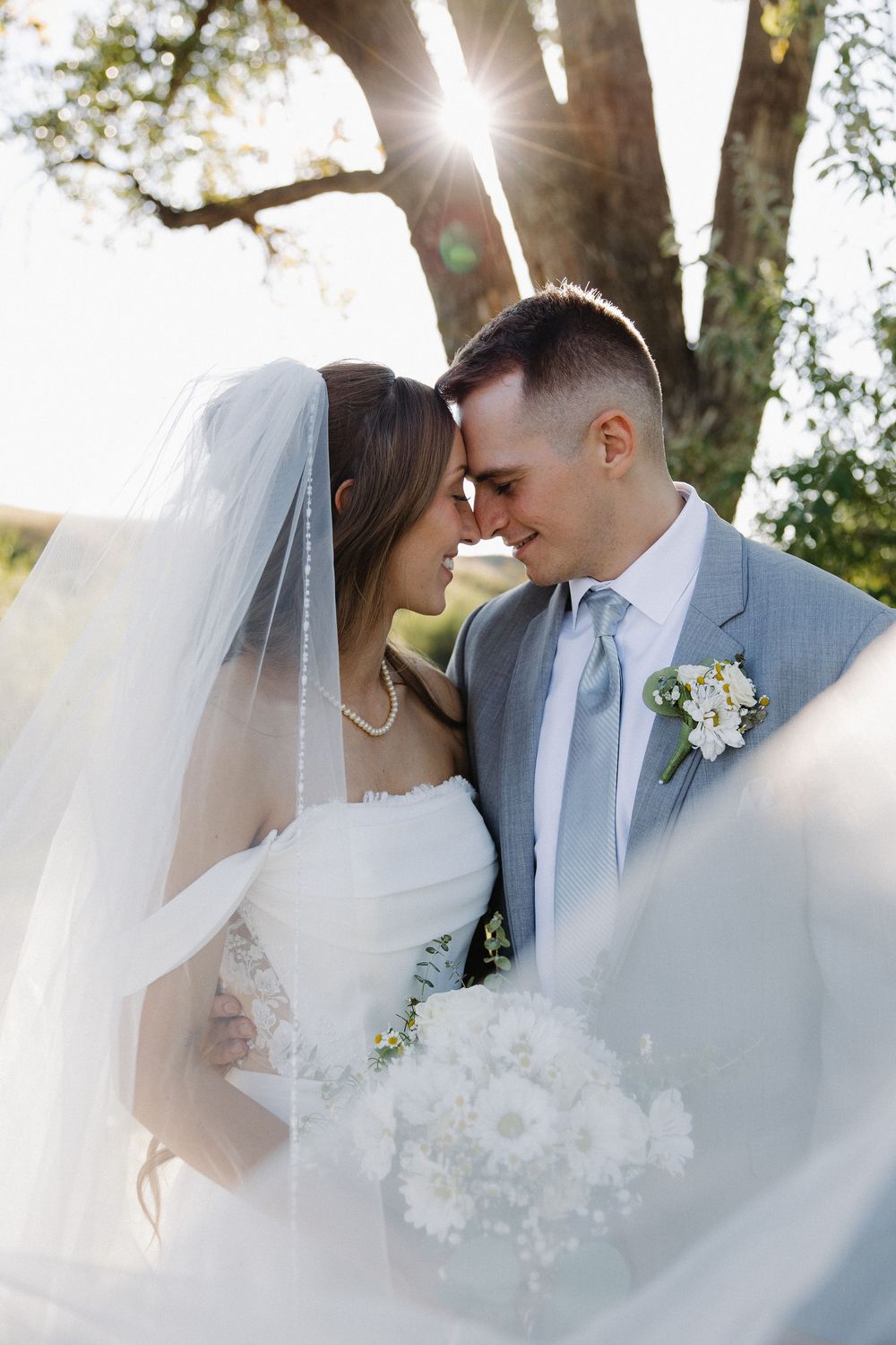 Bride and groom touch foreheads tenderly as veil billows in sunlit outdoor wedding portrait.
Great Falls Montana, Milo Creek Wedding photogr
