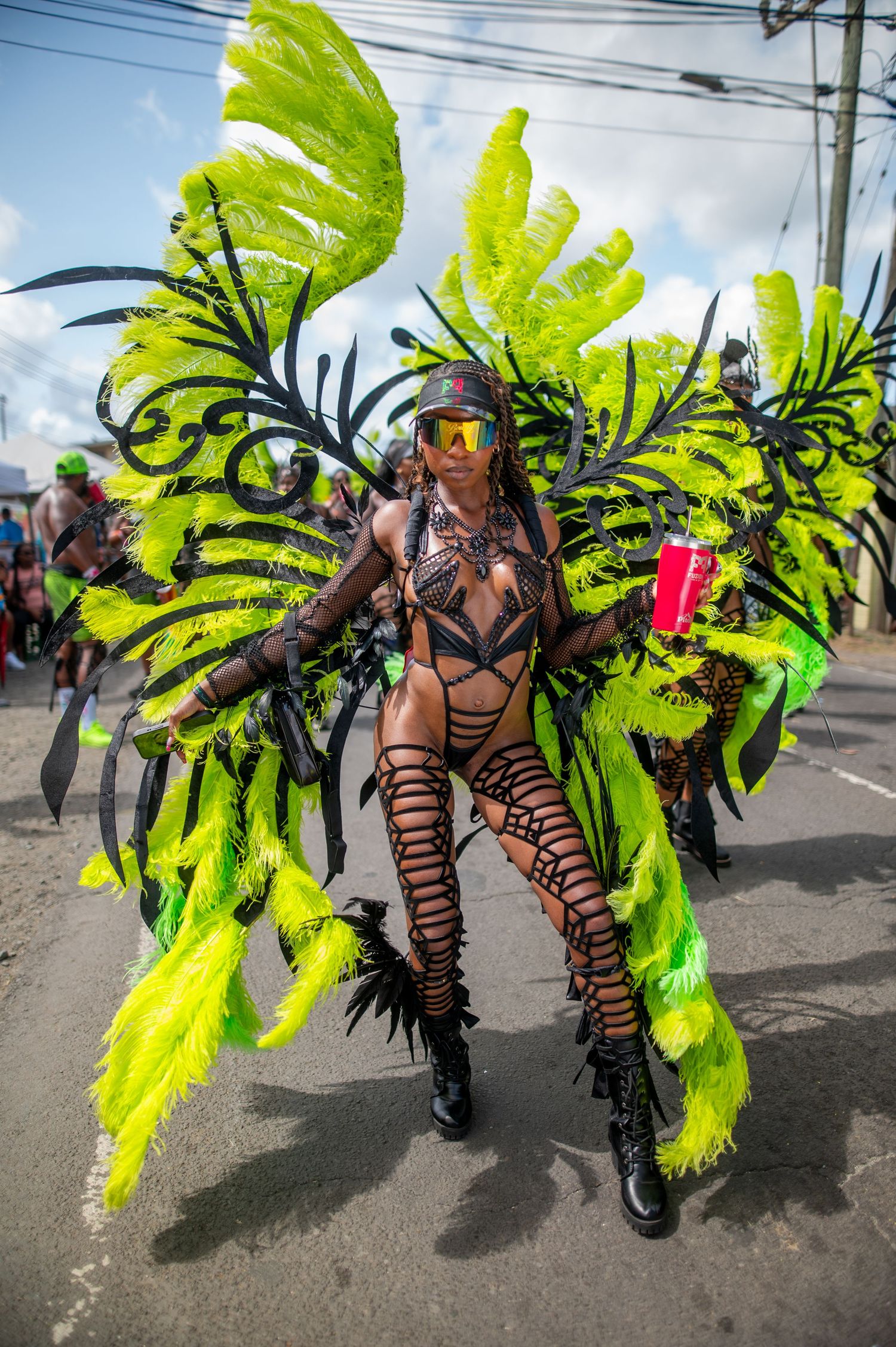 Carnival performer in vibrant neon green and black feathered costume with boots dancing in street parade.