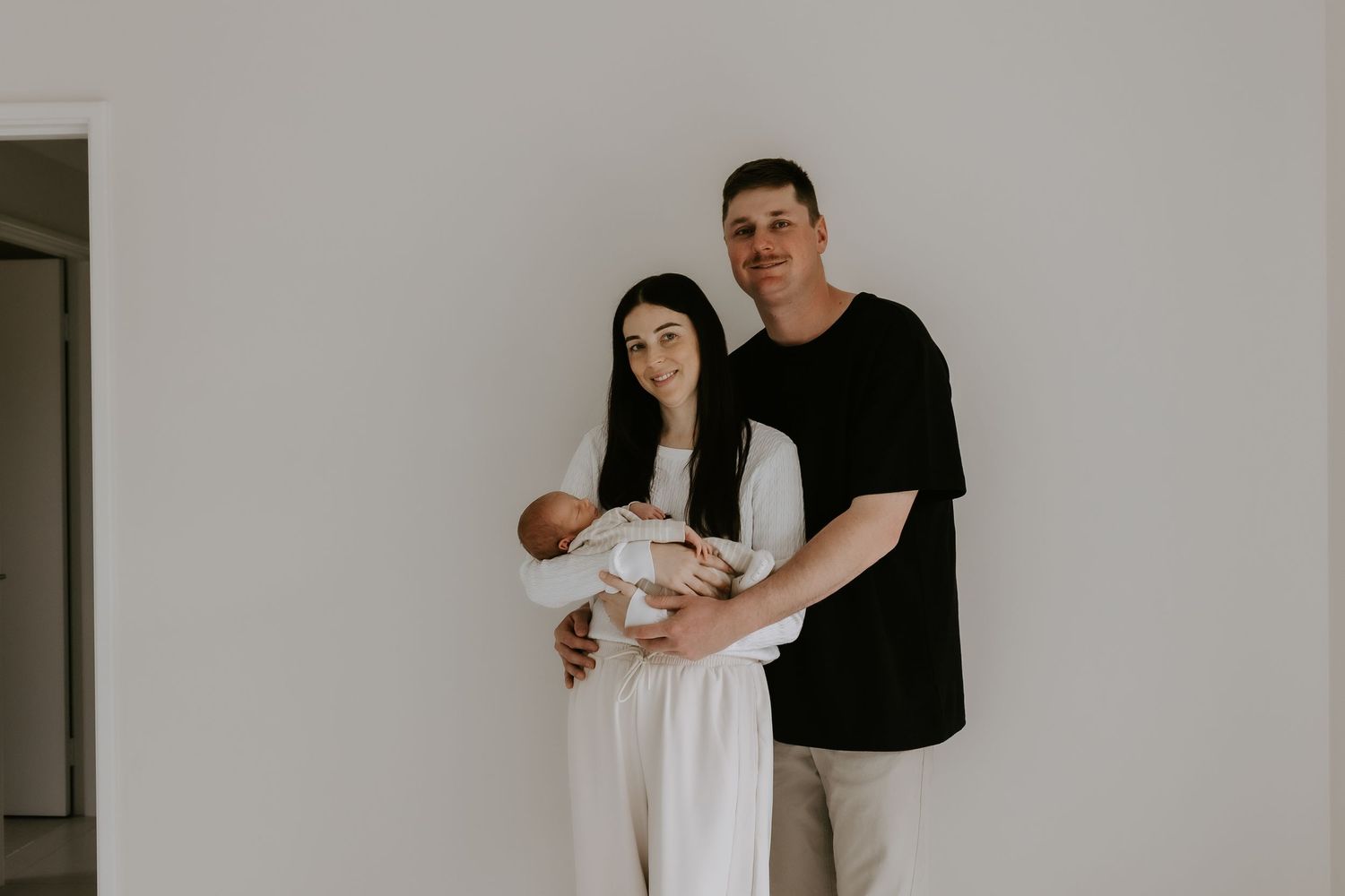 A young couple smiling and holding their newborn baby against a white wall in a minimalist family portrait.
