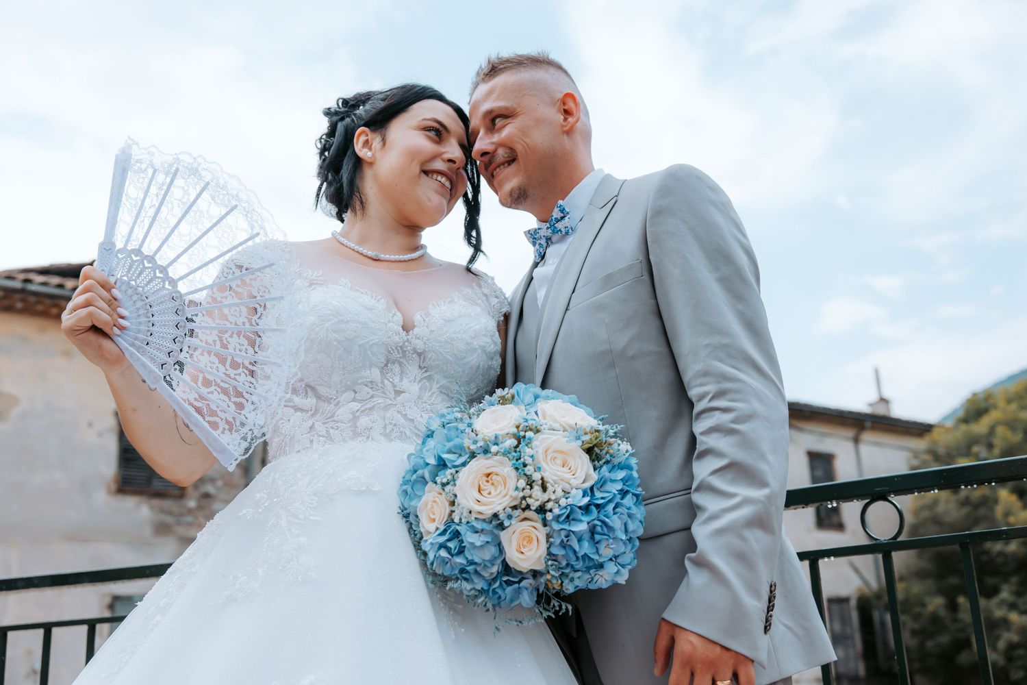 Smiling bride in white lace gown with tattoo holds blue bouquet beside groom in gray suit outdoors.