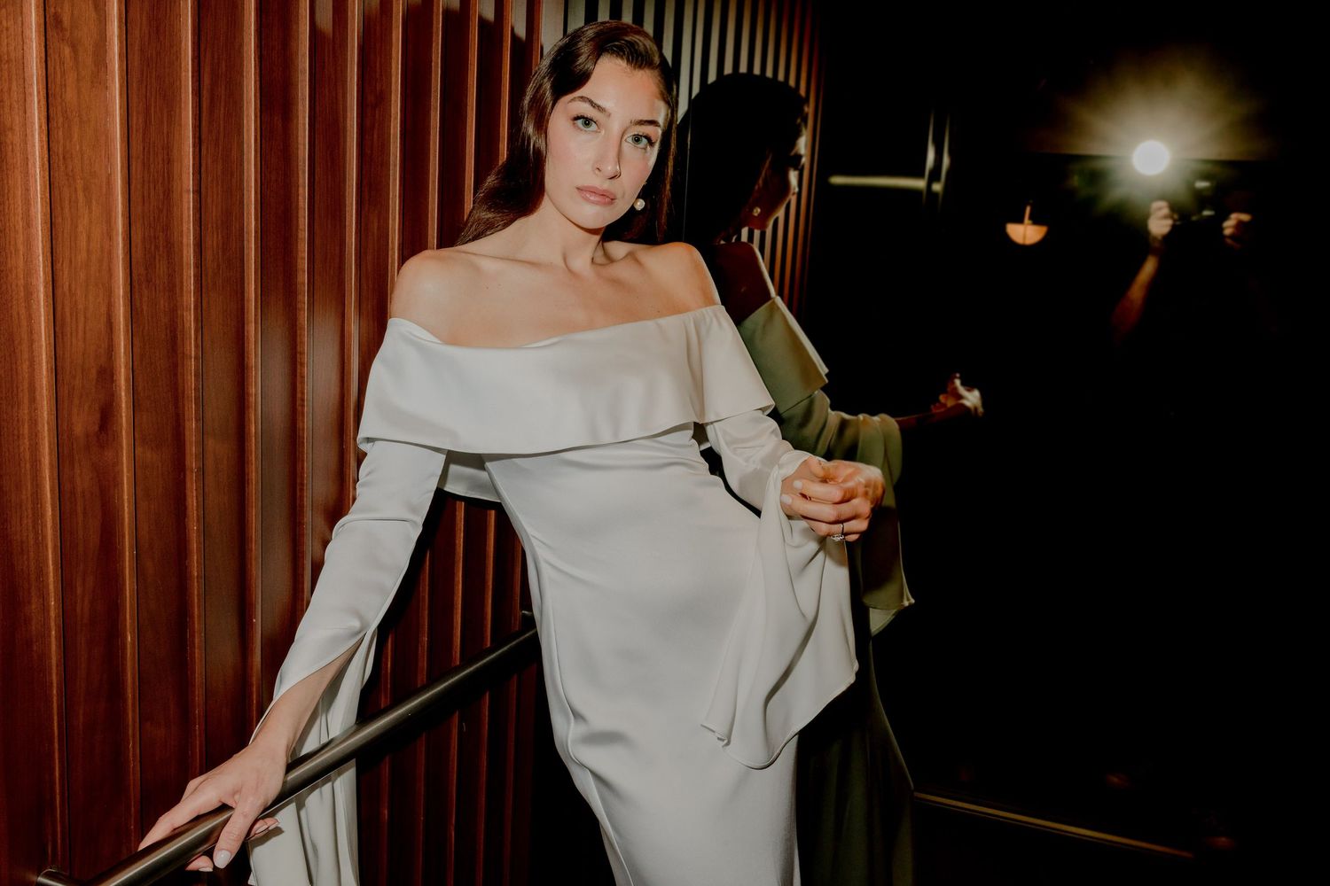 The Bride in elegant off-shoulder sage green gown posing on staircase in dimly lit venue.