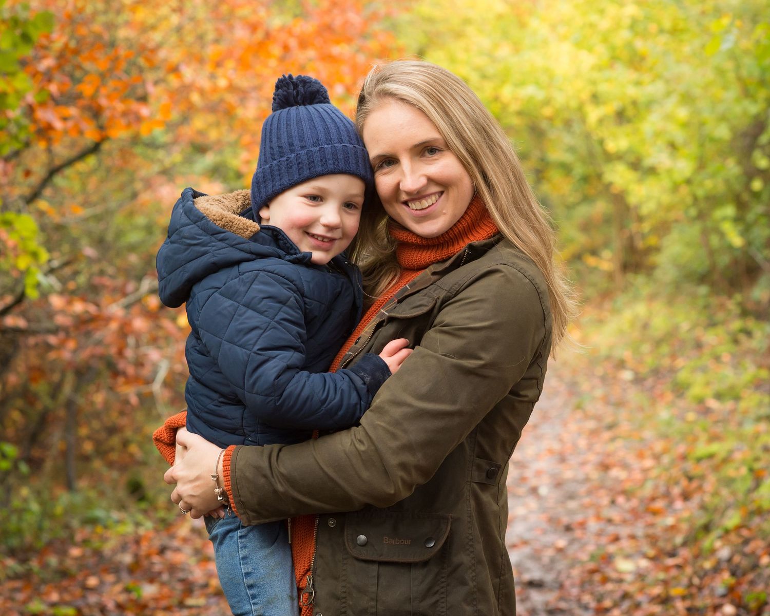 Smiling mother in an olive coat hugs a young child in a navy winter coat and knit hat on a leafy autumn forest path, warm and joyful.