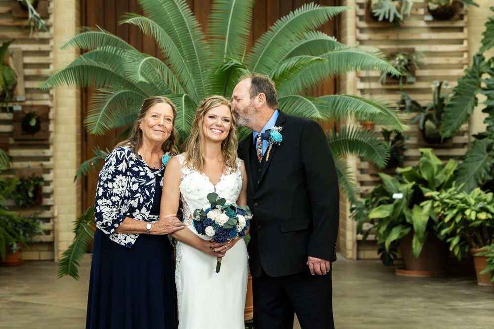 Bride in white gown holds bouquet flanked by family members in formal attire at an indoor garden wedding venue.