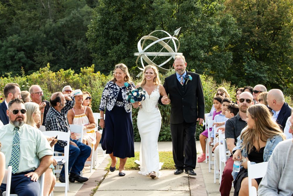 Bride walks down outdoor aisle with family, guests seated, lush greenery and armillary sphere backdrop.