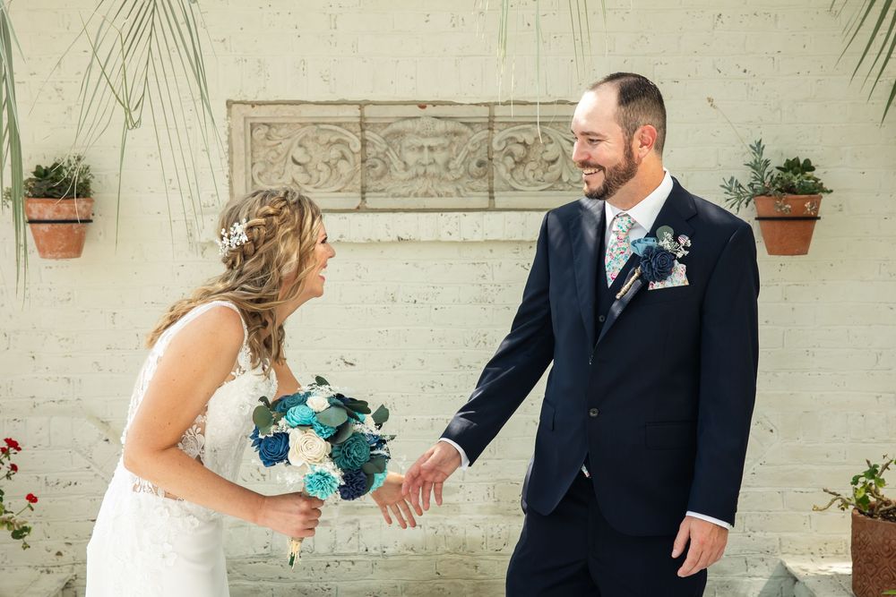 Bride and groom share a joyful first look moment, she holds a teal bouquet, he wears a navy suit with floral boutonniere.