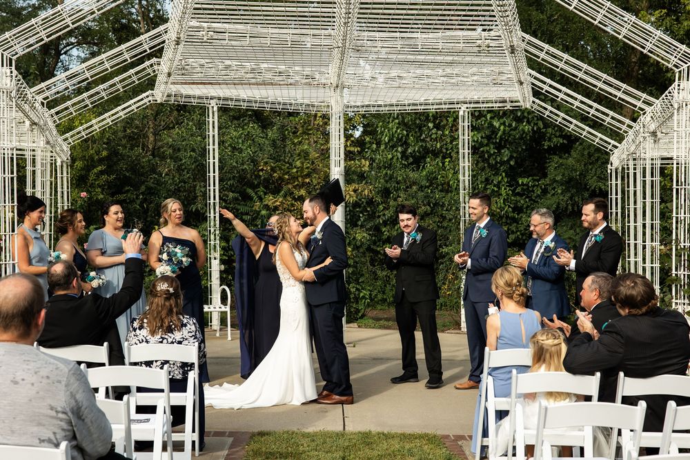 Couple shares first kiss at outdoor wedding ceremony under white geometric gazebo with wedding party and guests applauding.