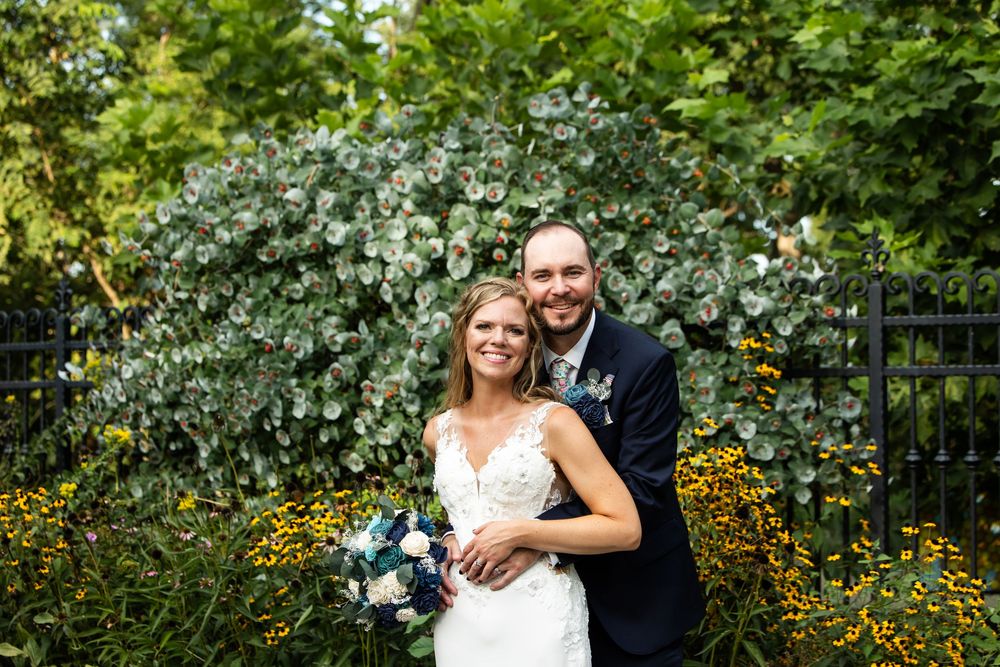 Happy couple poses in front of lush green bushes and yellow flowers on their wedding day.