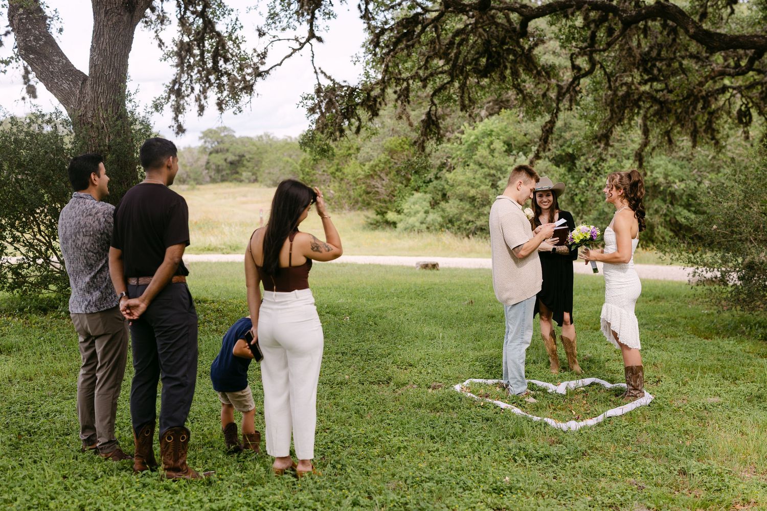 Small outdoor wedding ceremony under oak tree with guests gathered on green lawn.