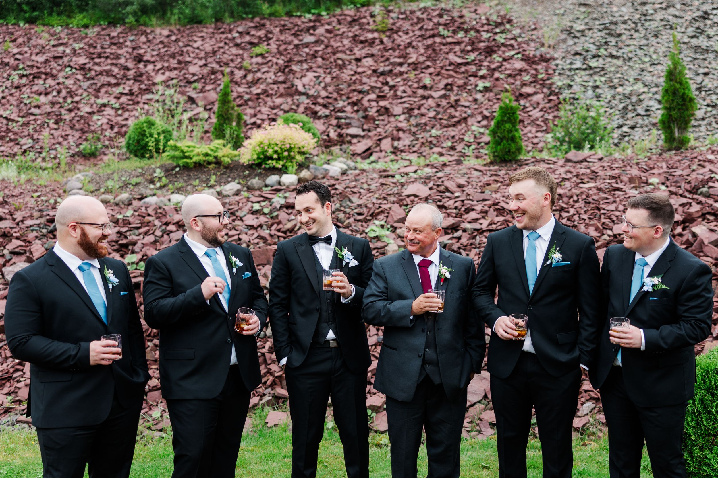 Six groomsmen in black suits with teal ties laughing and celebrating outdoors at a wedding reception.