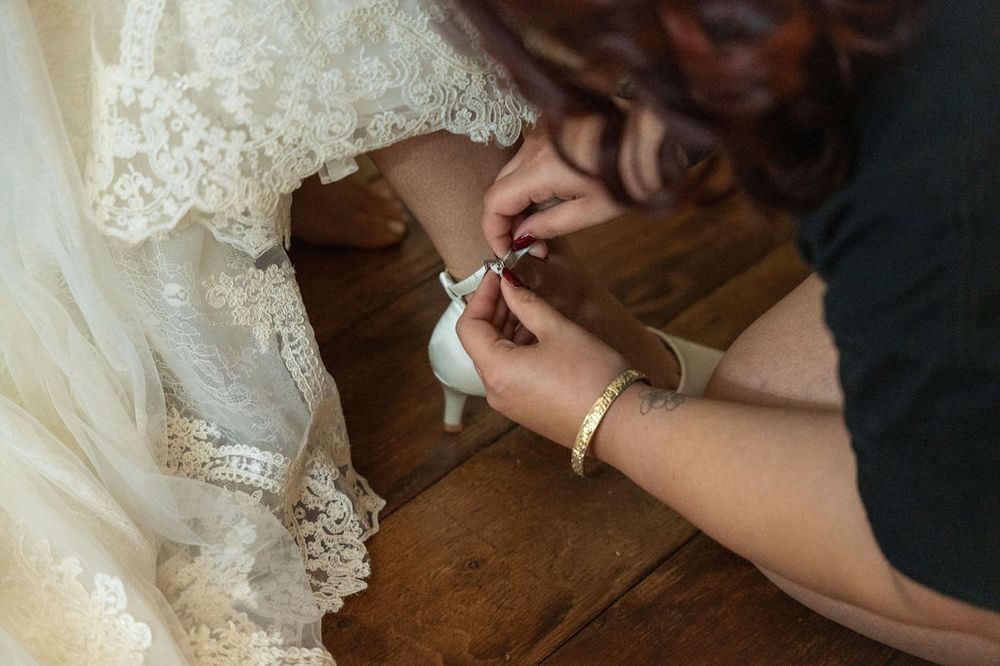 A bridesmaid fastening a white lace wedding dress button near the hem on a wooden floor.