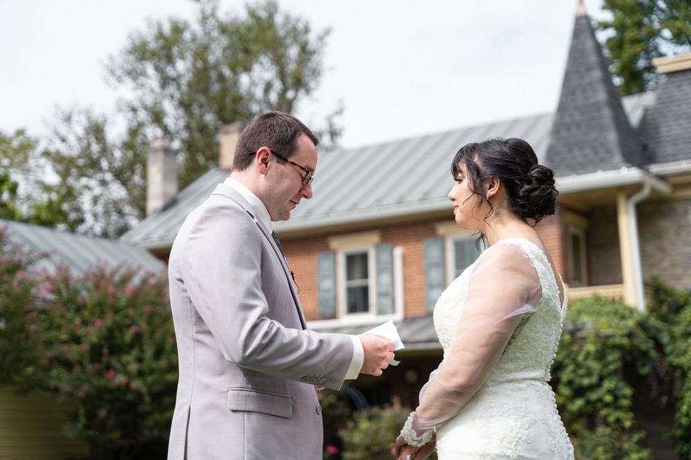 Bride and groom share a tender first look moment outdoors, holding hands in front of a charming brick estate.