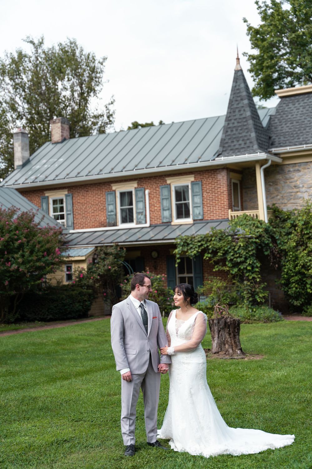 Bride and groom hold hands on green lawn before charming brick estate with blue shutters and metal roof.