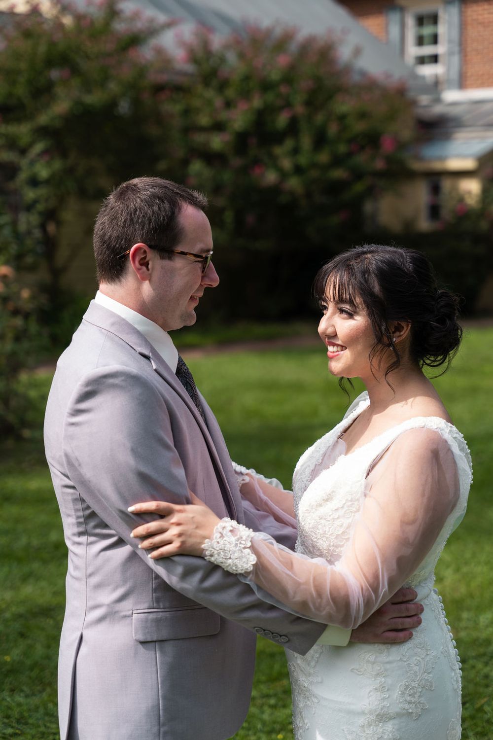 A bride and groom share a tender moment outdoors, smiling at each other in wedding attire on a green lawn.