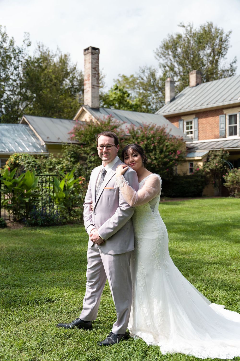 Bride and groom pose together on green lawn outside historic brick venue on their wedding day.