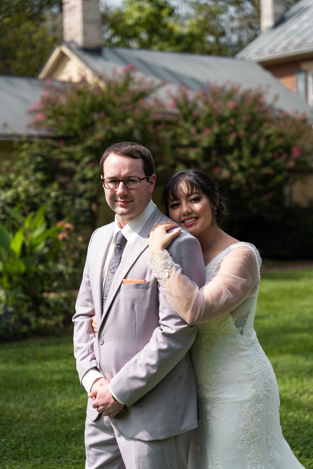 Bride and groom pose together outdoors on green lawn, she wears lace dress, he wears gray suit with tie.
