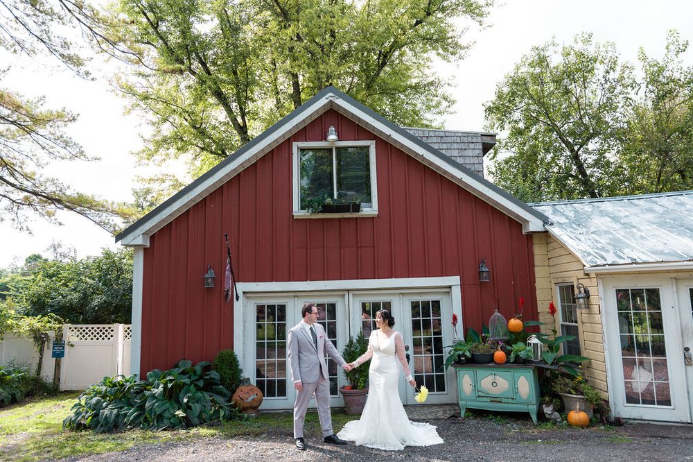 Bride and groom holding hands outside a red barn wedding venue surrounded by lush green trees and autumn decor.