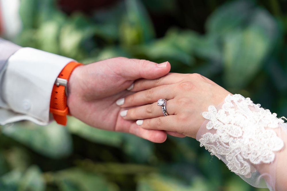 Close-up of bride and groom holding hands, showcasing diamond engagement ring and lace wedding dress sleeve.