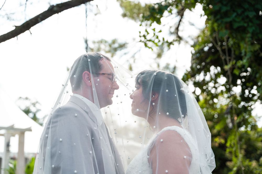 Bride and groom share a romantic moment under a dotted wedding veil outdoors surrounded by lush greenery.