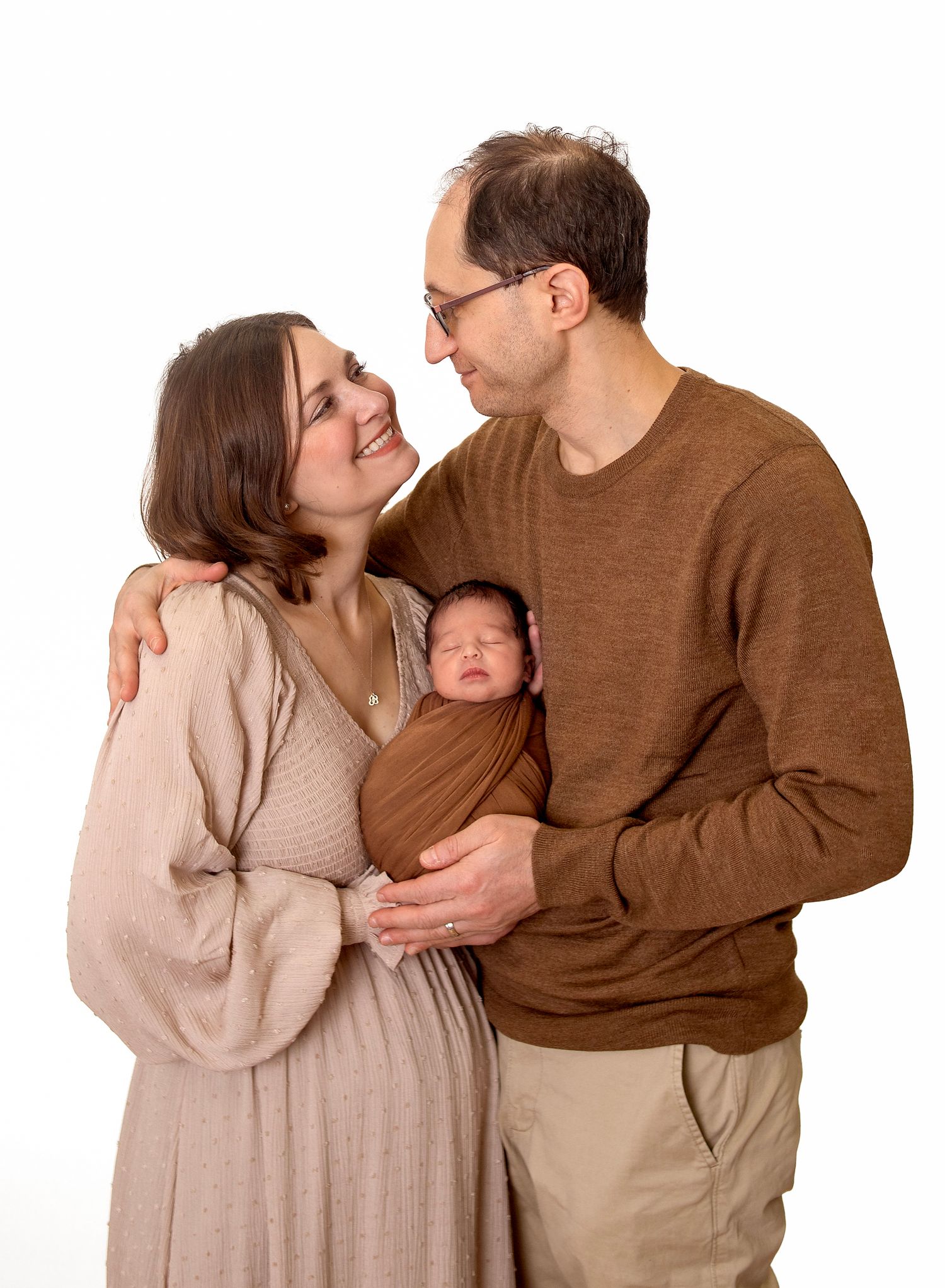 Happy couple smiling at each other while holding their sleeping newborn baby wrapped in a brown swaddle blanket.