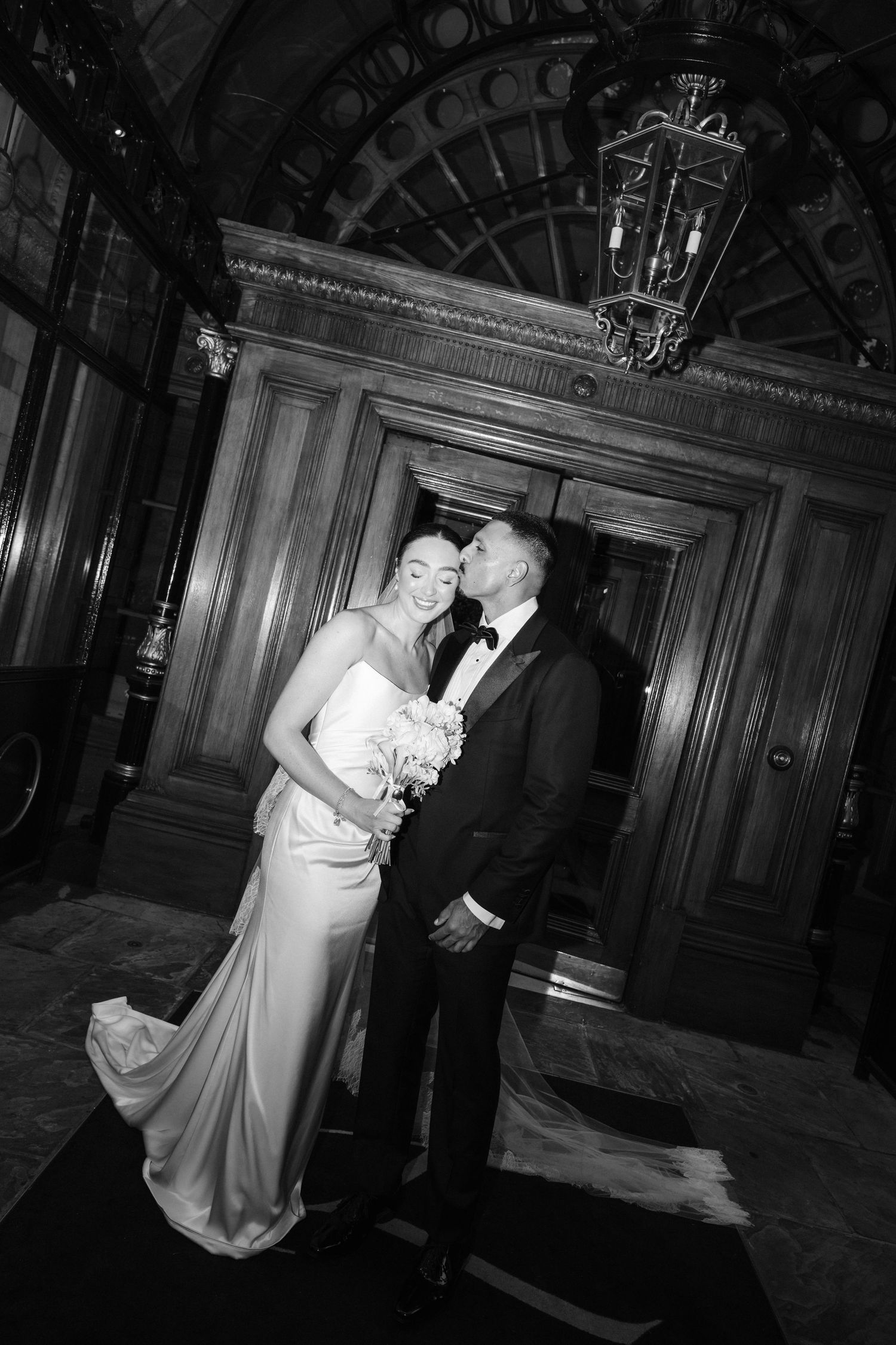 Black and white photo of a bride and groom sharing a romantic kiss in an elegant wood-paneled venue.