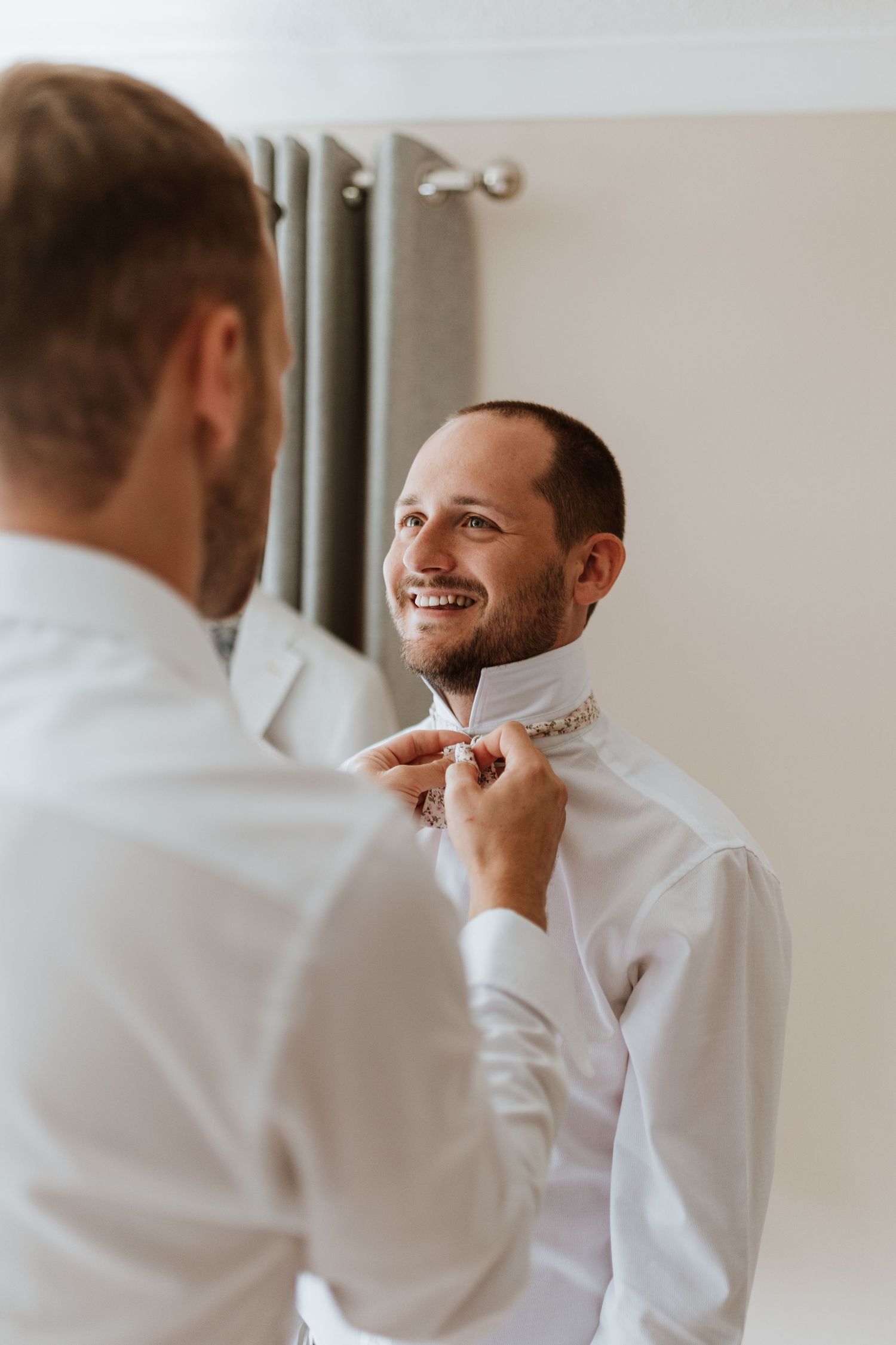 Two grooms in white dress shirts smiling while one helps the other button his collar before a wedding ceremony.