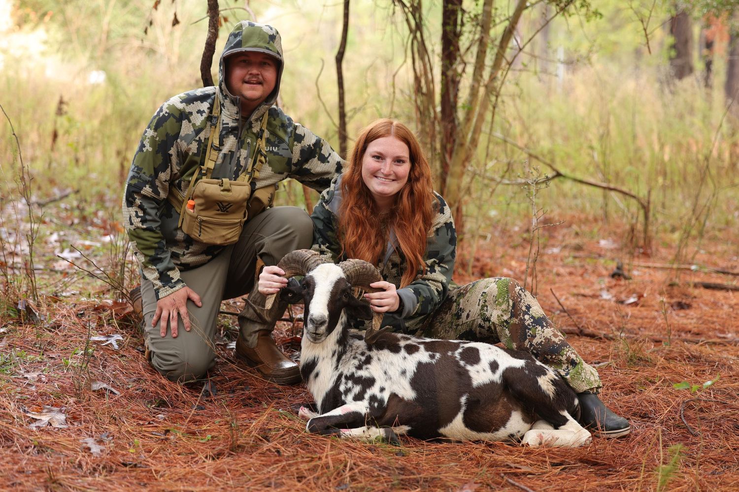Two hunters in camouflage posing with harvested goat in wooded hunting area.