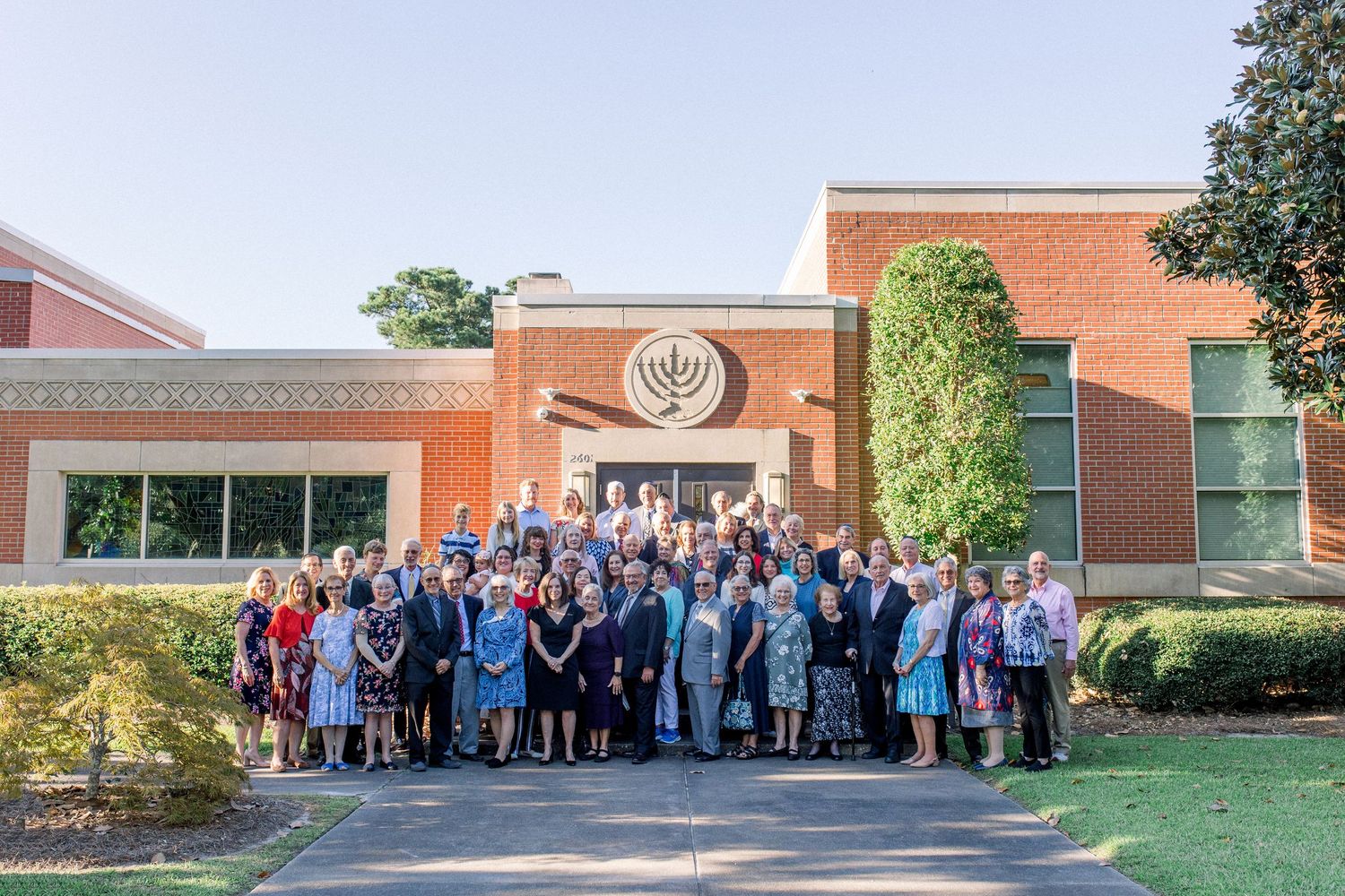 Large group of people posing outside a brick synagogue building with a menorah emblem, on a sunny day.